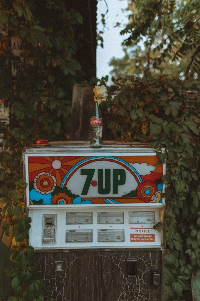 Colorful retro 7-Up vending machine adorned with vintage floral designs, surrounded by lush greenery.